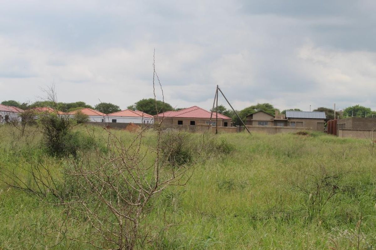 A view of a vacant residential plot featuring natural grass cover with surrounding residential properties in the background.