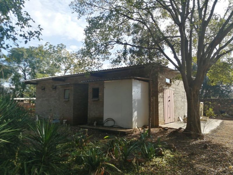 A rear view of a rustic farm building featuring exposed brick walls, a corrugated metal roof, and mature surrounding trees.
