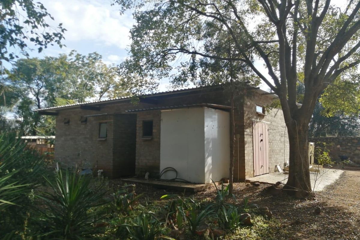 A rear view of a rustic farm building featuring exposed brick walls, a corrugated metal roof, and mature surrounding trees.