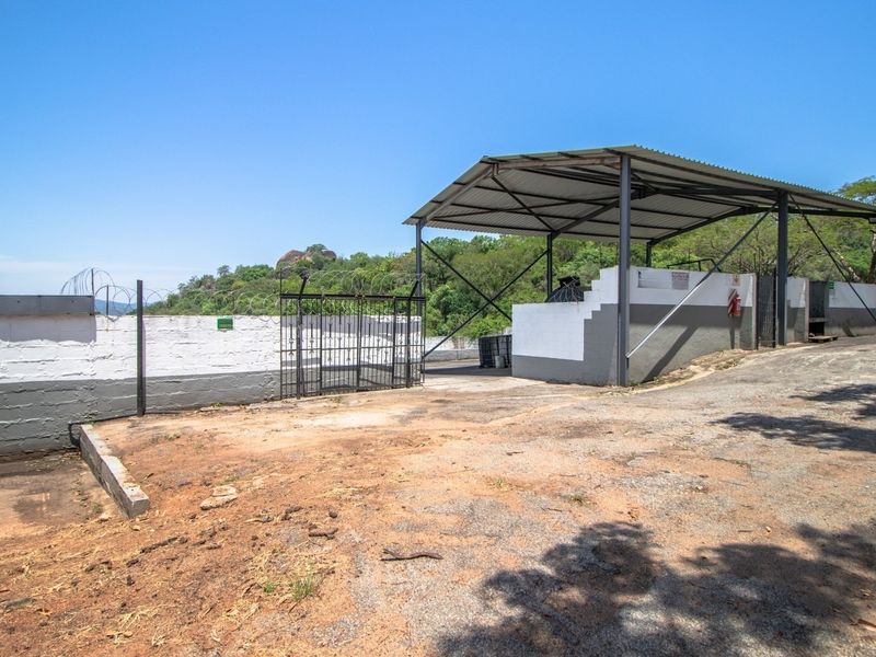 An outdoor utility area featuring a covered metal structure, perimeter wall with security fencing, and a concrete driveway on a farm property.