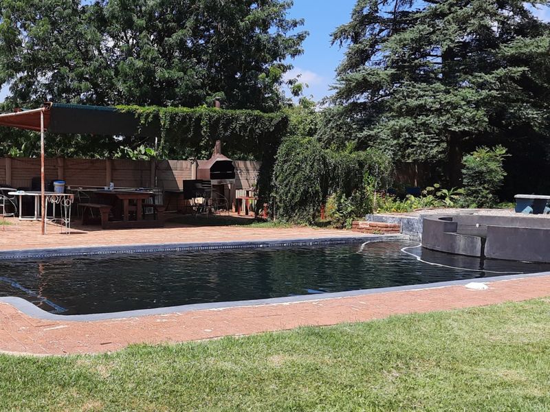 Outdoor pool area featuring a swimming pool, paved patio, built-in braai, and a shaded entertainment space set within a garden.