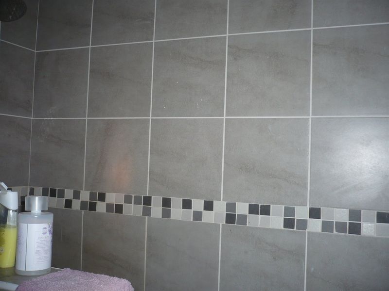 A close-up view of a bathroom shower area featuring large format grey wall tiles with a decorative mosaic accent border.