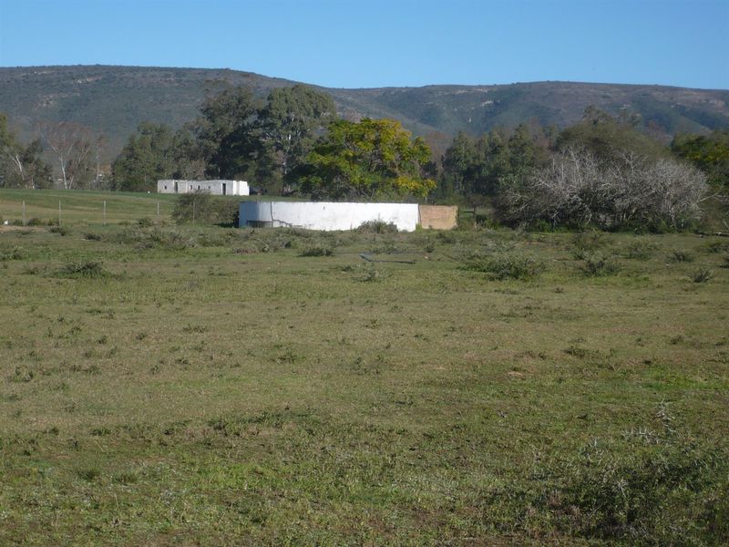 A wide landscape view of the farm property featuring an open field, a circular water reservoir, and distant outbuildings against a mountain backdrop.