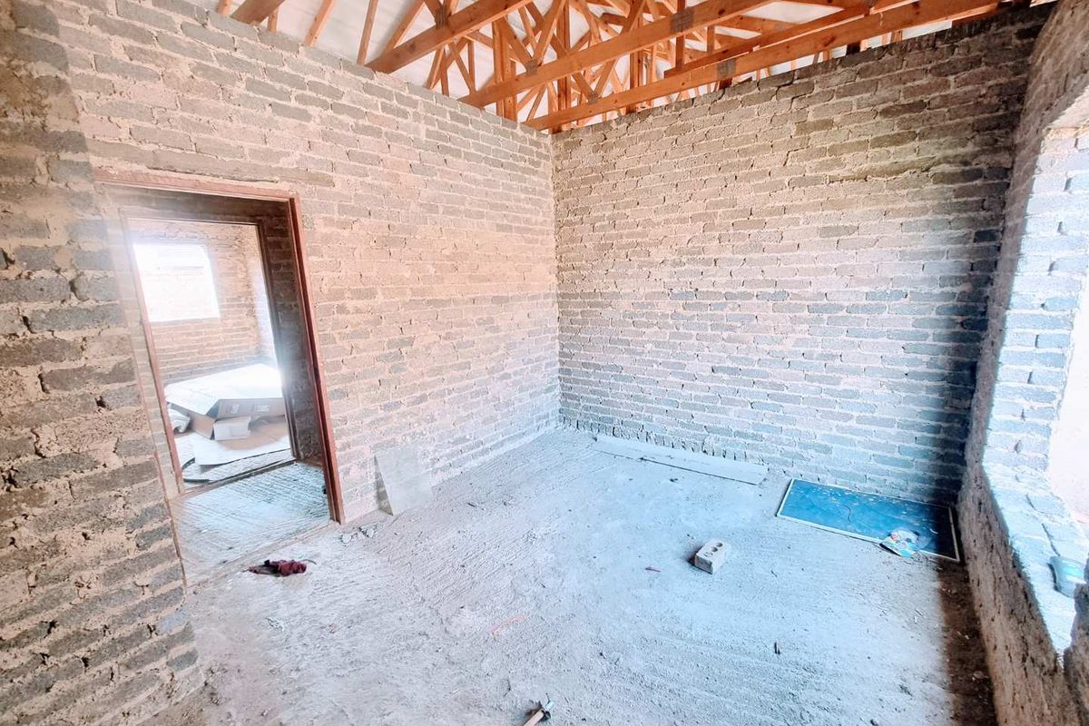 An unfinished interior room featuring exposed brick walls, open roof trusses, and raw concrete flooring, currently in the construction phase.