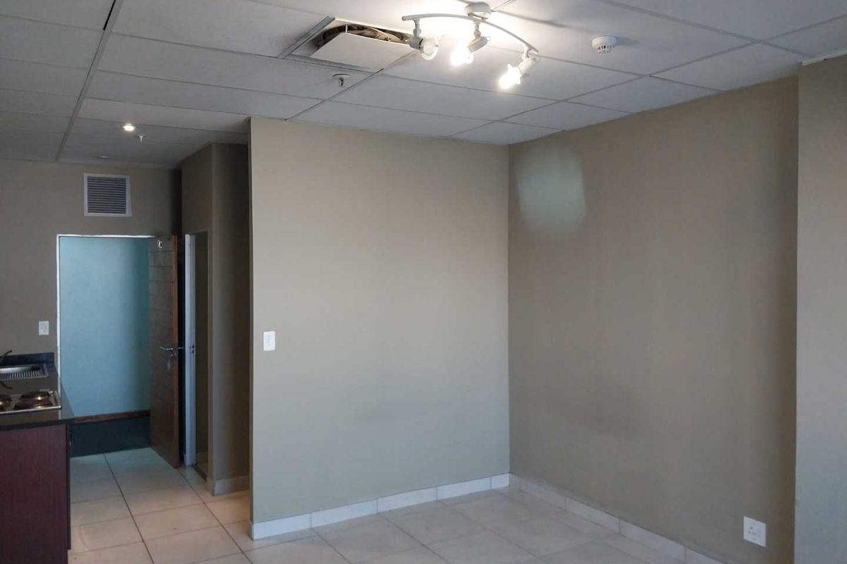 An empty living area featuring tiled flooring, a drop ceiling with a missing tile, and a partial view into an adjacent kitchen space.