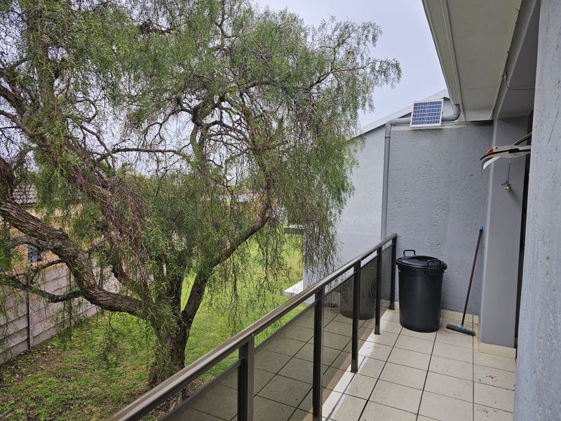 A balcony featuring tiled flooring and a glass balustrade, overlooking a garden area with a large tree.