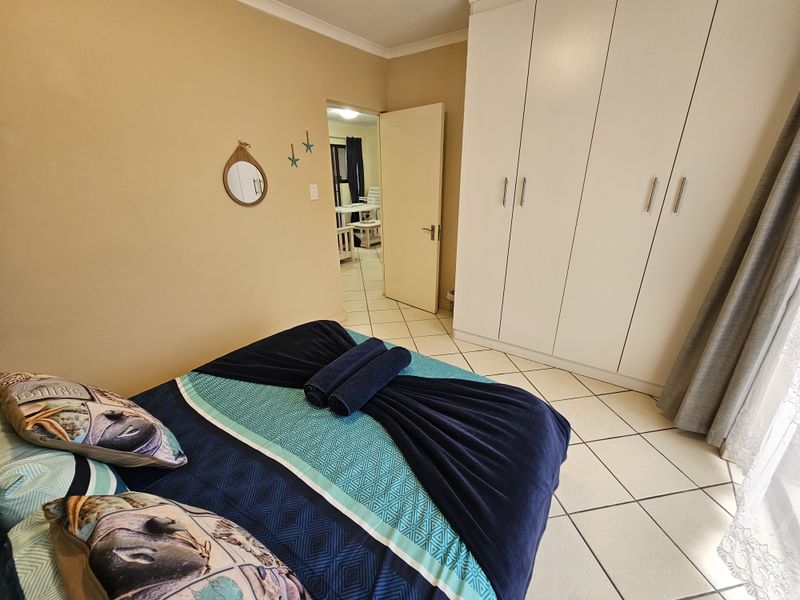 A bedroom featuring tiled flooring, built-in white cupboards, and neutral wall tones, with a doorway leading to an adjacent living area.