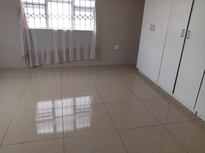 A clean, empty bedroom featuring neutral tiled flooring, built-in white cupboards, and windows fitted with security bars.