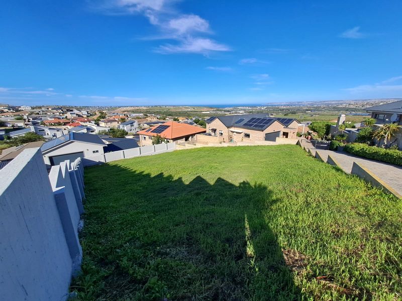A vacant residential plot featuring a sloped grassy area, boundary walls, and a paved driveway with expansive views of the surrounding neighborhood and coastline.
