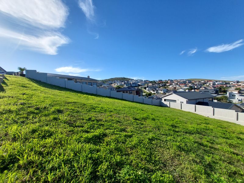 A view of a sloped, vacant residential plot featuring a perimeter boundary wall and surrounding neighborhood views.