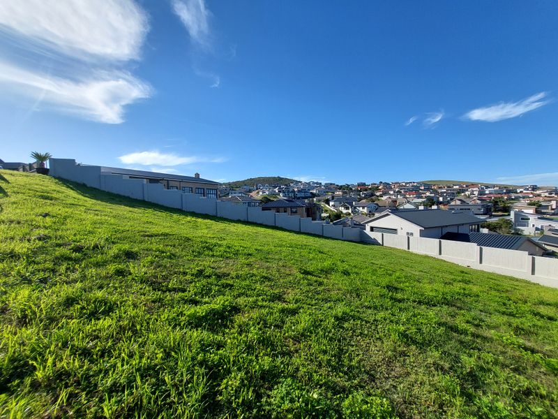 A view of a sloped residential vacant land plot featuring a boundary wall and surrounding neighborhood scenery.