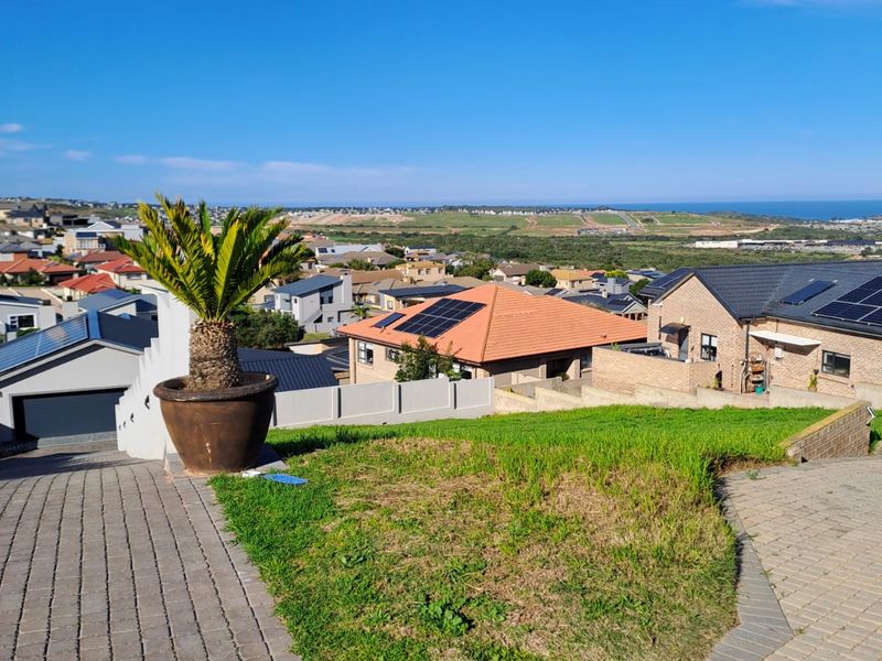 Elevated view overlooking a residential neighborhood with coastal scenery in the distance, featuring paved driveway access and established landscaping.