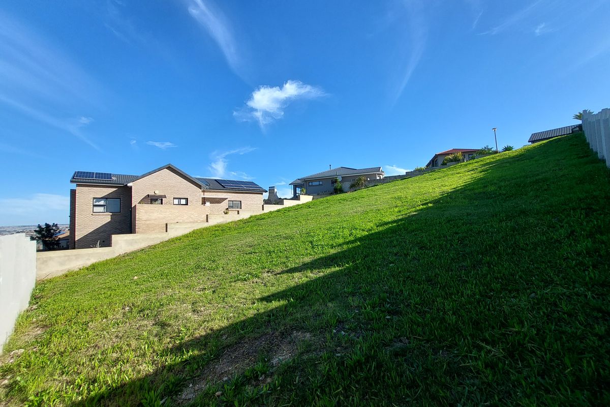 A view of a sloped residential plot featuring a grass lawn, retaining wall, and neighboring houses with solar panels installed on the roofs.