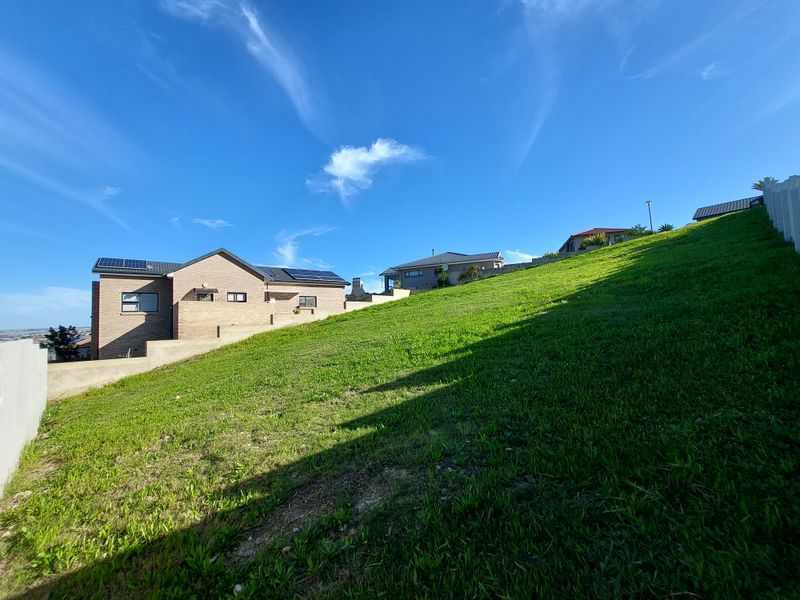 A view of a sloped residential plot featuring a grass lawn, retaining wall, and neighboring houses with solar panels installed on the roofs.