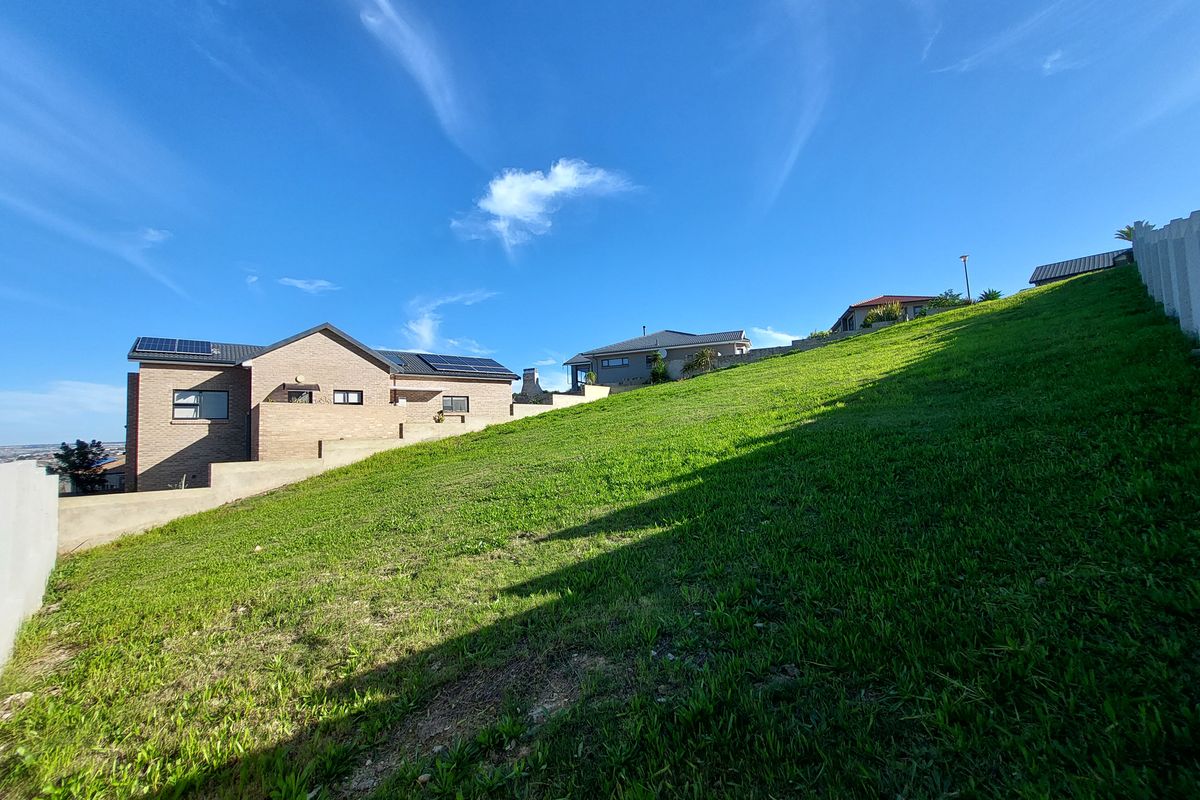 A view of a sloped residential plot featuring a well-maintained grass lawn, boundary walls, and neighboring properties in the background.