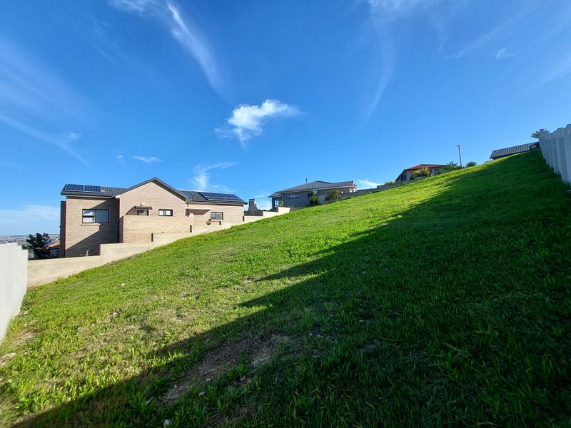 A view of a sloped residential plot featuring a well-maintained grass lawn, boundary walls, and neighboring properties in the background.