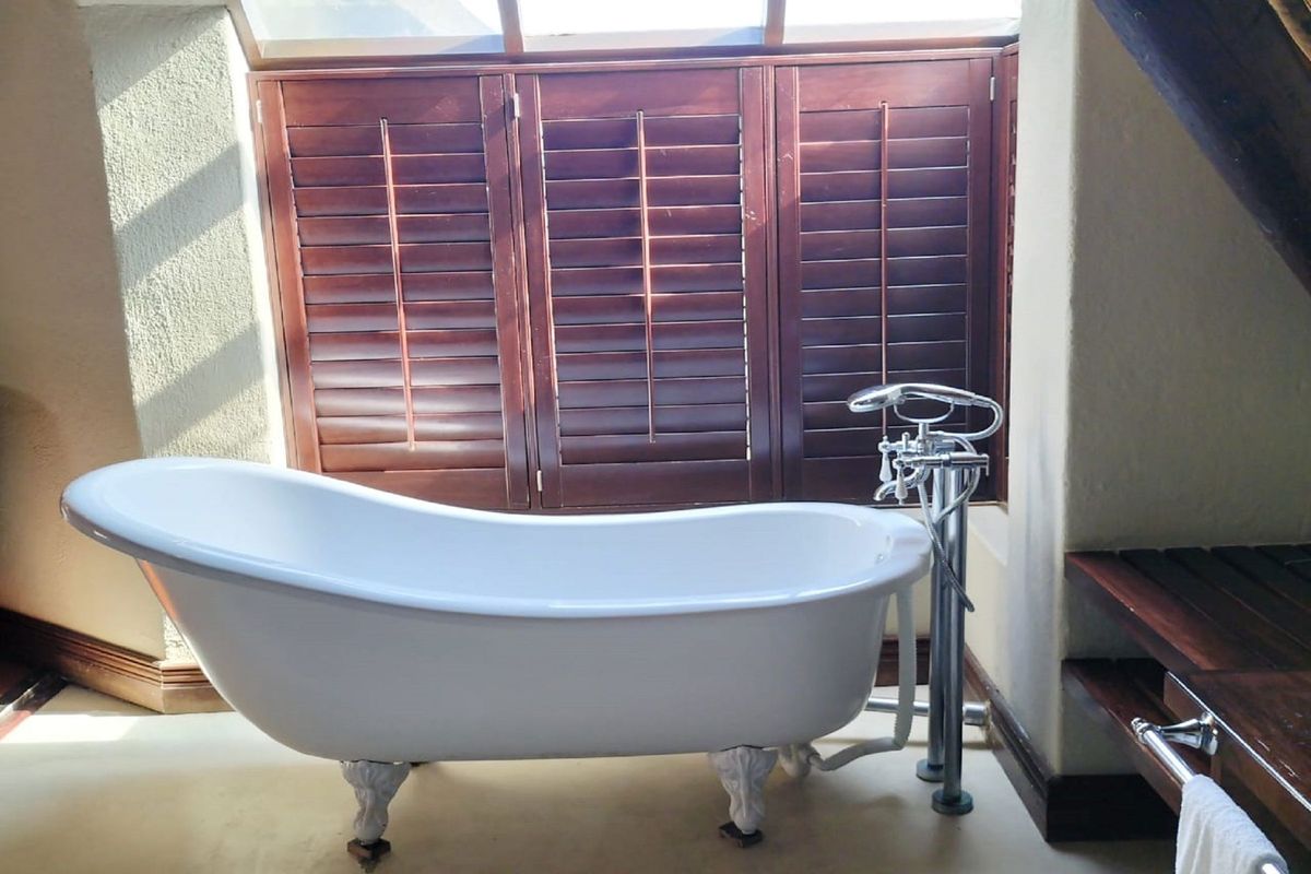 A bright bathroom featuring a classic white clawfoot bathtub, floor-mounted chrome faucet, and elegant wooden plantation shutters under a skylight.