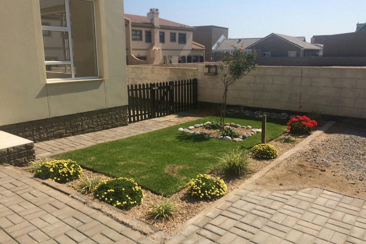 A rear garden area featuring a paved patio, a small lawn with landscaped borders, and a boundary wall with a timber gate.