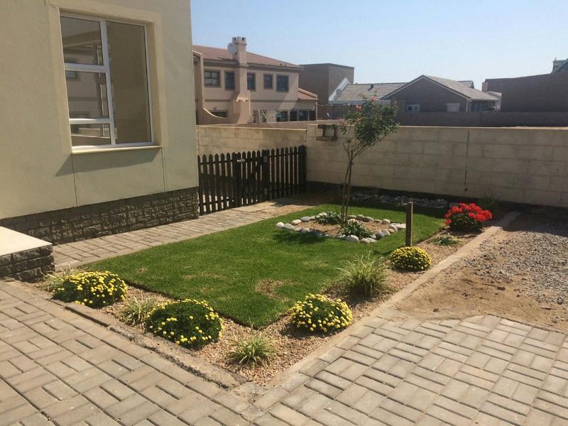 A rear garden area featuring a paved patio, a small lawn with landscaped borders, and a boundary wall with a timber gate.