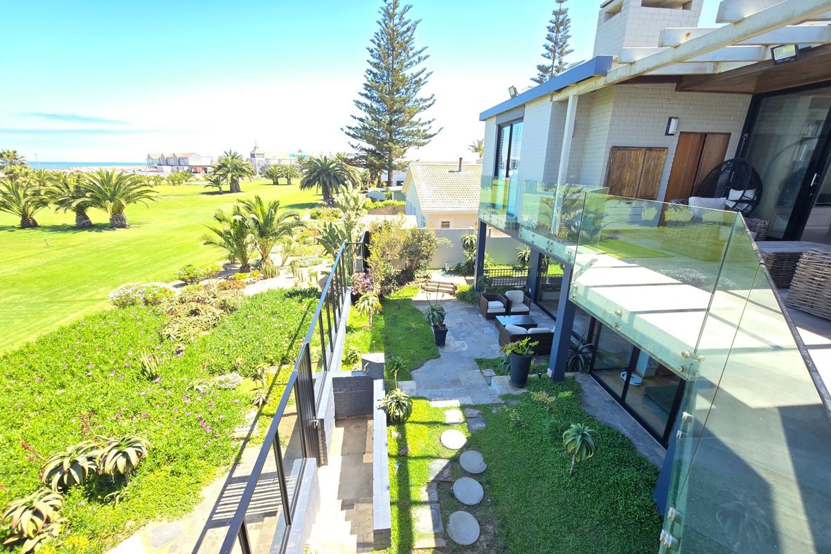 Elevated view of a modern rear exterior featuring a stone patio, glass balustrade, and a well-maintained garden overlooking a coastal landscape.