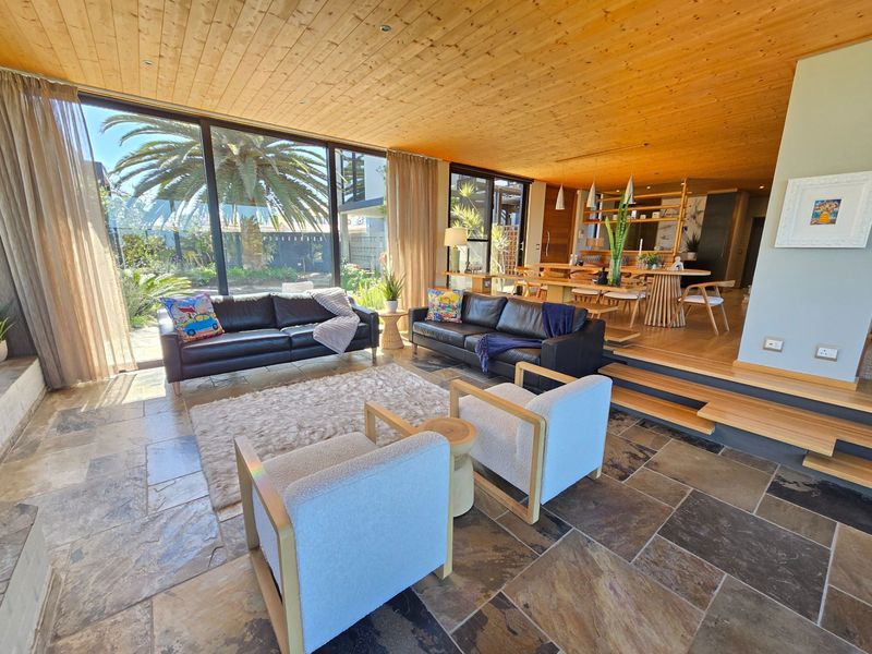 Spacious open-plan living and dining area featuring a warm timber-clad ceiling, natural stone flooring, and expansive floor-to-ceiling glass doors opening to the garden.