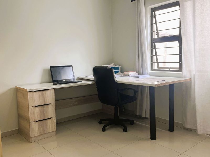 A functional home office space featuring tiled flooring, a built-in desk, and a window fitted with security bars.