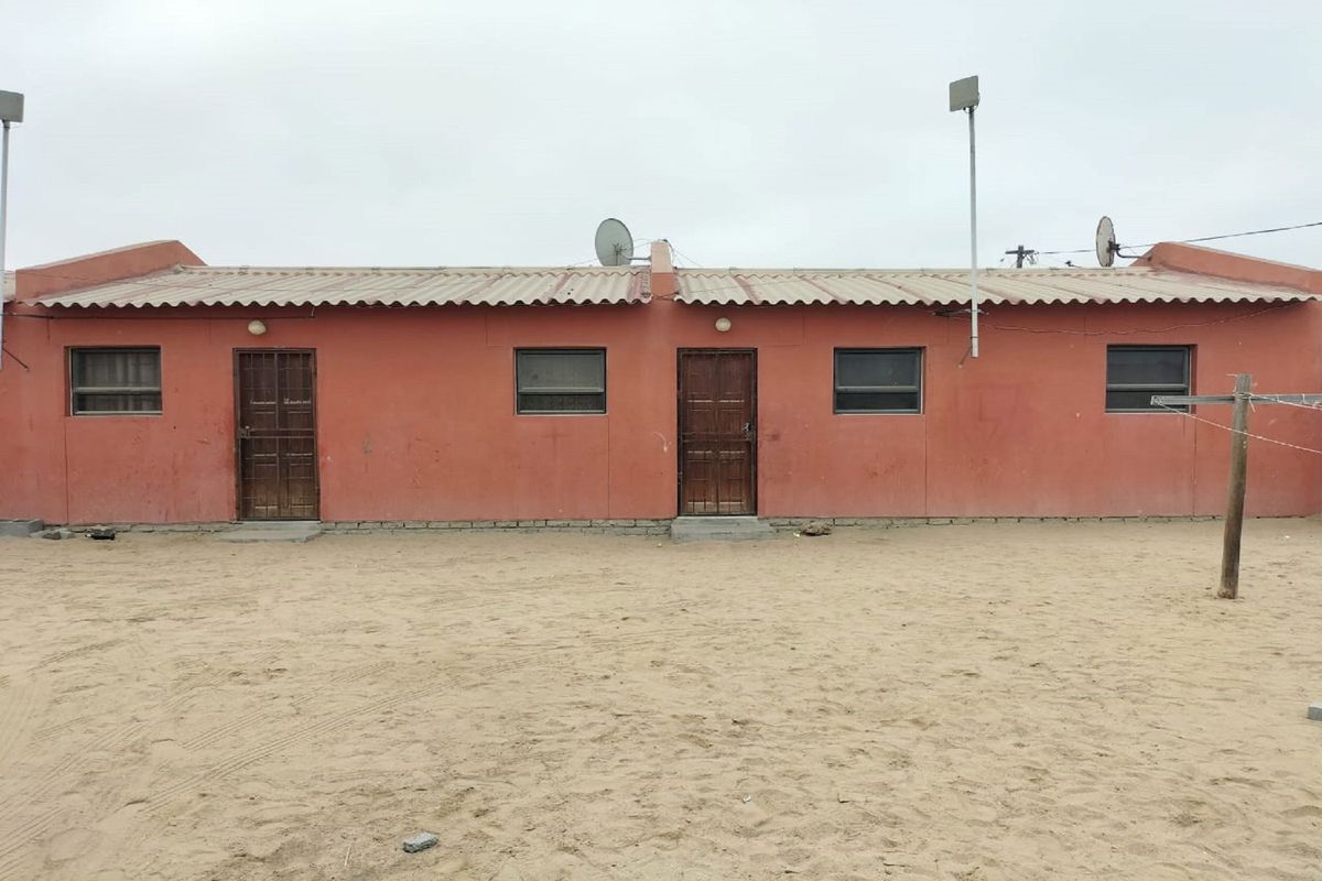 Rear exterior view of a single-story residential building featuring a corrugated roof, security doors, and a sandy yard area.