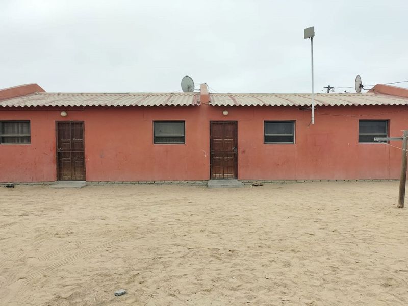 Rear exterior view of a single-story residential building featuring a corrugated roof, security doors, and a sandy yard area.