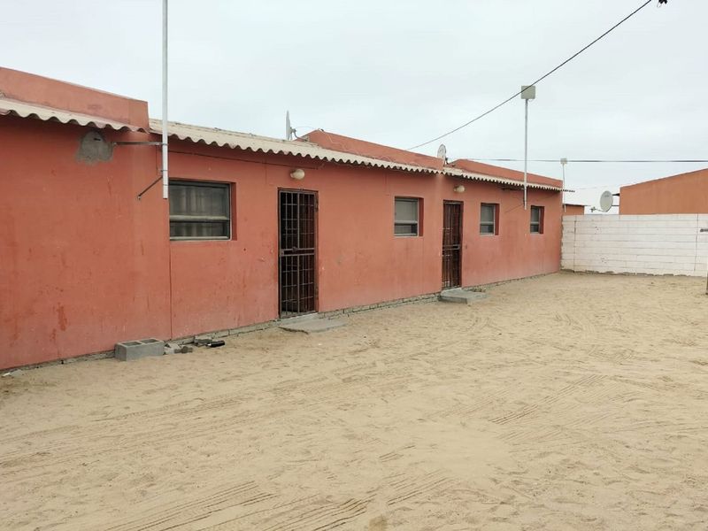 Rear view of a single-story residential property featuring a sandy yard, security gates, and a corrugated roof structure.