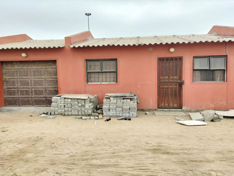 Front elevation of a single-story house featuring a garage, security gate, and corrugated roofing, situated on an unpaved lot.