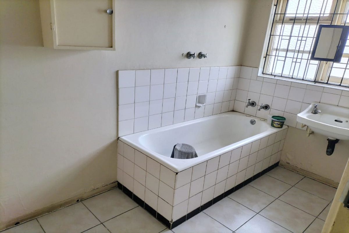 A dated bathroom featuring a tiled bathtub, wall-mounted basin, and security bars on the window, requiring cosmetic updates to improve its overall condition.