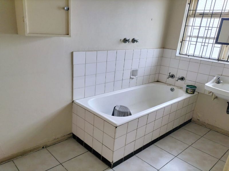 A dated bathroom featuring a tiled bathtub, wall-mounted basin, and security bars on the window, requiring cosmetic updates to improve its overall condition.