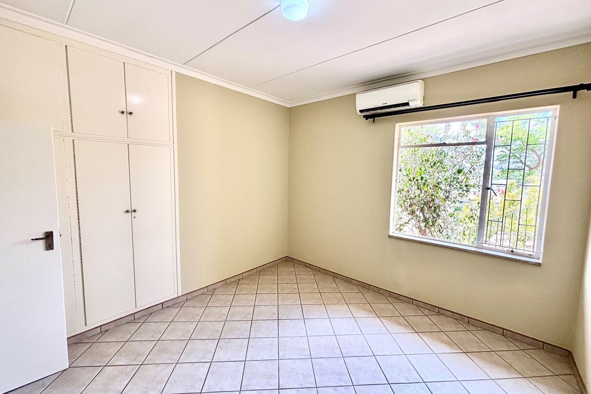 A clean, empty bedroom featuring tiled flooring, built-in cupboards, and an installed air conditioning unit with security bars on the window.