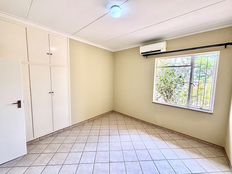 A clean, empty bedroom featuring tiled flooring, built-in cupboards, and an installed air conditioning unit with security bars on the window.