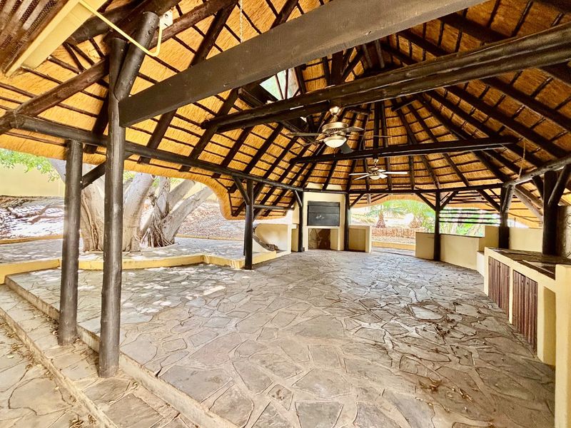 Spacious outdoor entertainment area featuring a thatched roof, exposed timber beams, stone flooring, and a built-in braai facility.