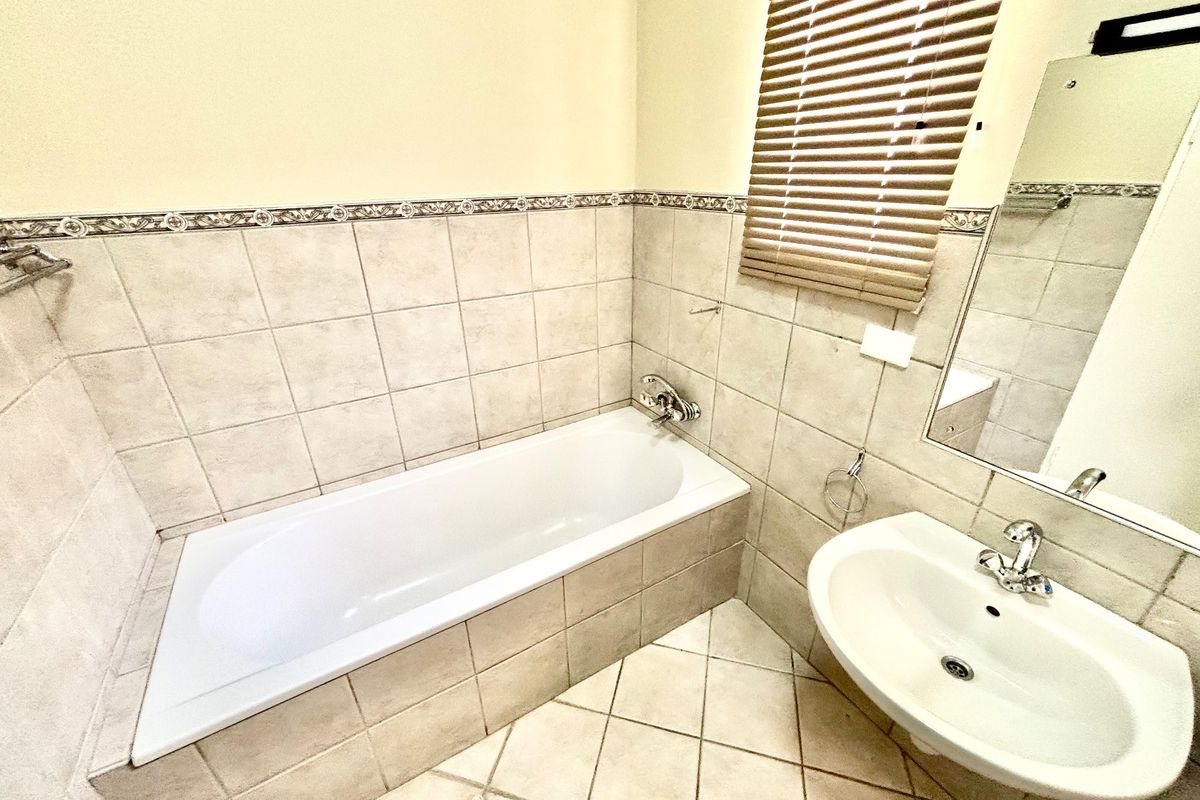 A functional bathroom featuring a built-in bathtub, wall-mounted basin, neutral ceramic tiling, and a wall mirror with integrated lighting.
