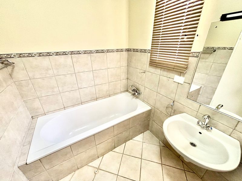 A functional bathroom featuring a built-in bathtub, wall-mounted basin, neutral ceramic tiling, and a wall mirror with integrated lighting.