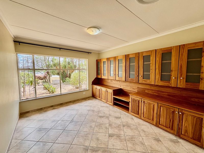 A bright, empty living space featuring extensive built-in wooden cabinetry, large windows with garden views, and neutral tiled flooring.
