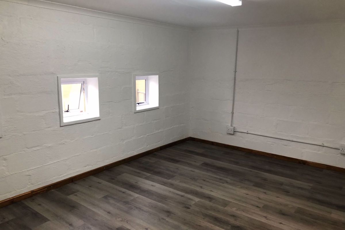 A clean, empty office space featuring modern laminate flooring, white painted block walls, and functional casement windows with exposed electrical conduit.