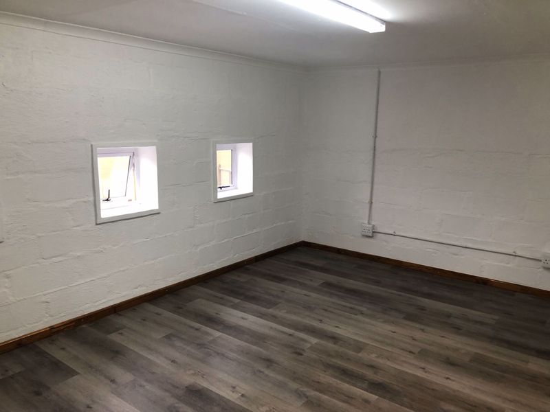 A clean, empty office space featuring modern laminate flooring, white painted block walls, and functional casement windows with exposed electrical conduit.