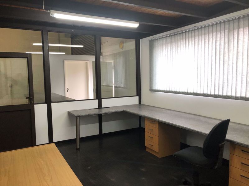 Functional office space featuring a built-in L-shaped desk, glass partition walls, and exposed ceiling beams, providing a professional industrial work environment.