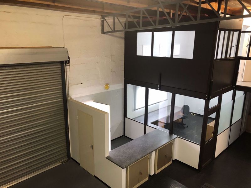 Industrial warehouse space featuring a roller shutter door, exposed timber roof trusses, and a multi-level partitioned office structure with built-in cabinetry.
