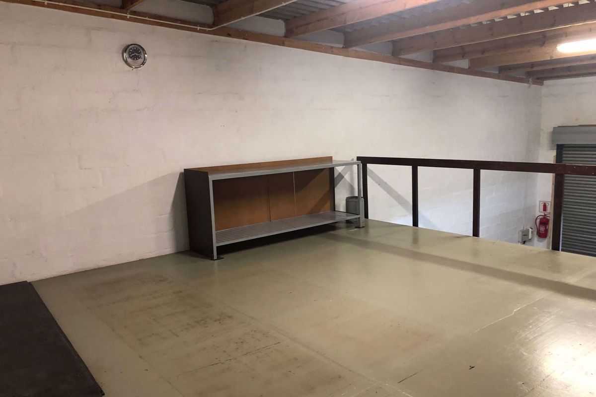 An open-plan industrial mezzanine level featuring exposed timber beams, metal flooring, safety railing, and a roller shutter door access point.