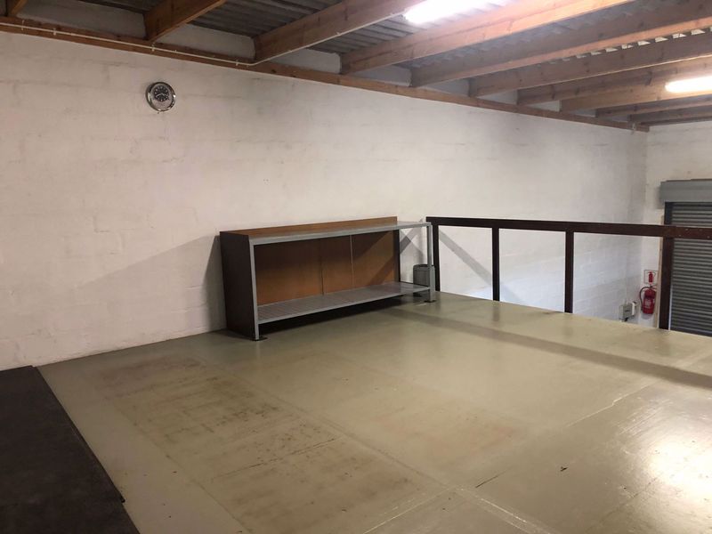 An open-plan industrial mezzanine level featuring exposed timber beams, metal flooring, safety railing, and a roller shutter door access point.