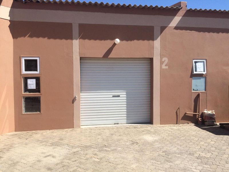 Front facade of an industrial unit featuring a secure roller shutter door, paved driveway access, and functional exterior lighting.