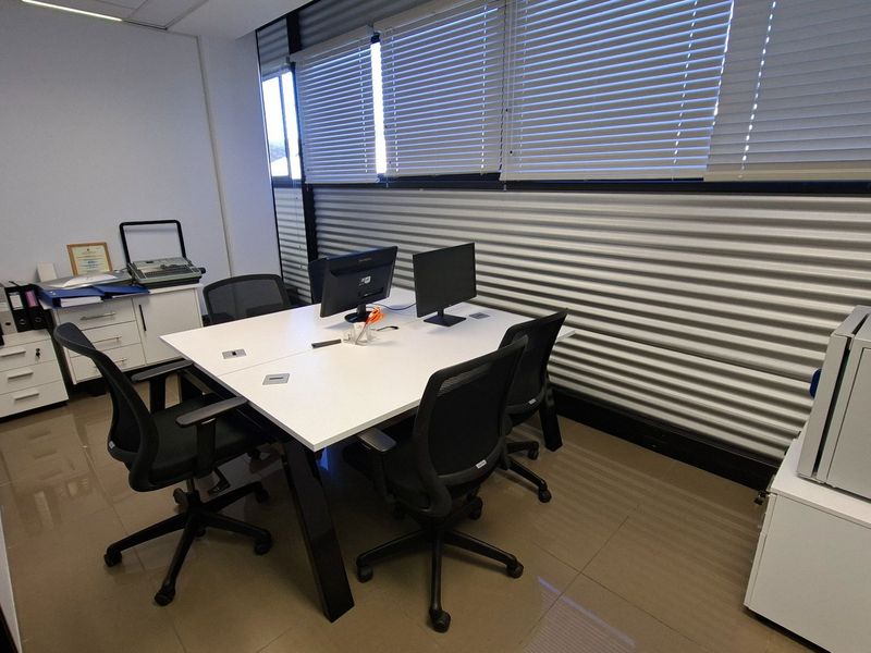 A functional office space featuring tiled flooring, a suspended ceiling, and large windows with adjustable venetian blinds for natural light control.