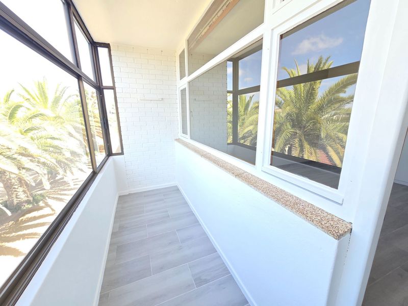 A bright, enclosed balcony featuring large windows, modern grey tiled flooring, and a clean white painted brick finish.