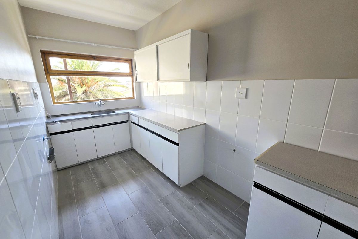 A clean, empty kitchen featuring white built-in cabinetry, laminate countertops, a stainless steel sink, and modern grey tiled flooring.