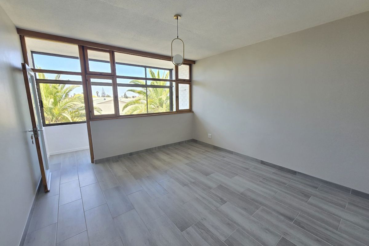 A bright, empty living area featuring modern wood-look tile flooring, large timber-framed windows, and a contemporary pendant light fixture.
