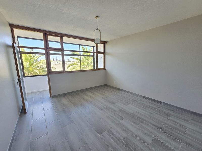A bright, empty living area featuring modern wood-look tile flooring, large timber-framed windows, and a contemporary pendant light fixture.